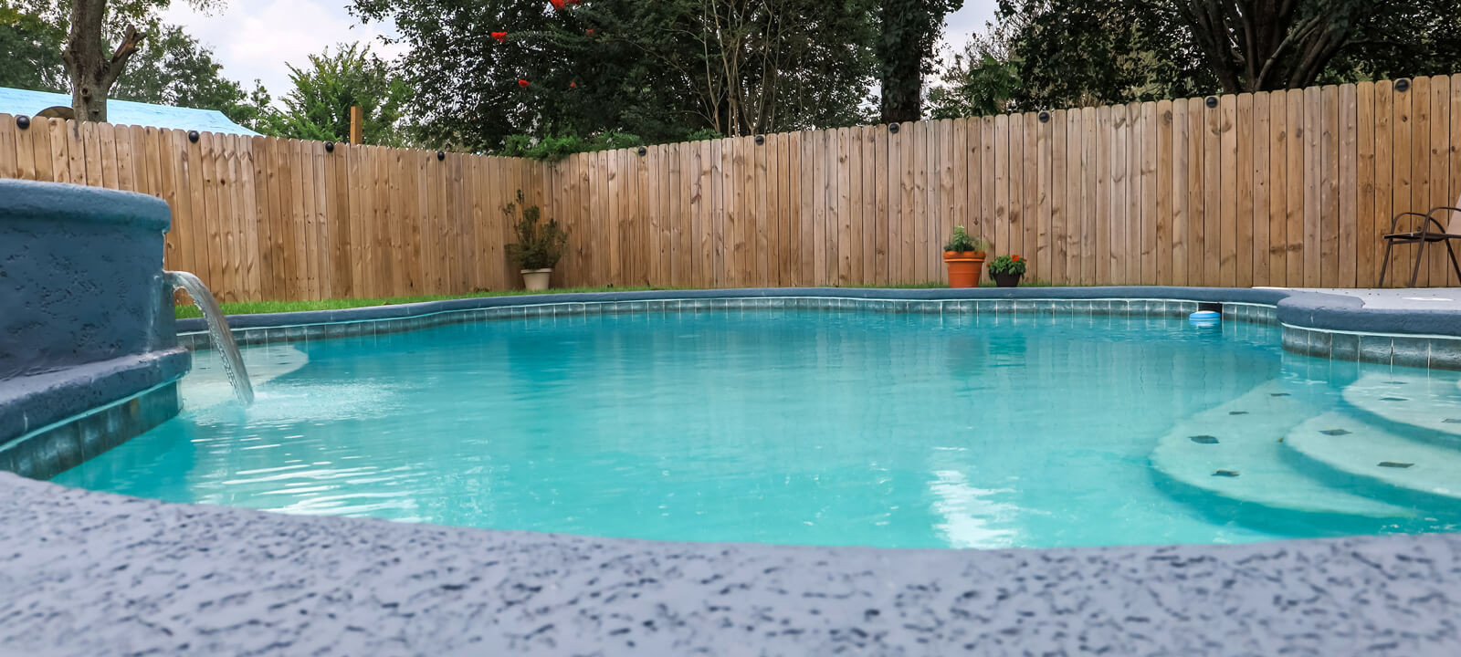 Low Angle View Of A Large Free Form Gray Grey Accent Swimming Pool With Turquoise Blue Water In A Fenced In Backyard In A Suburban Neighborhood